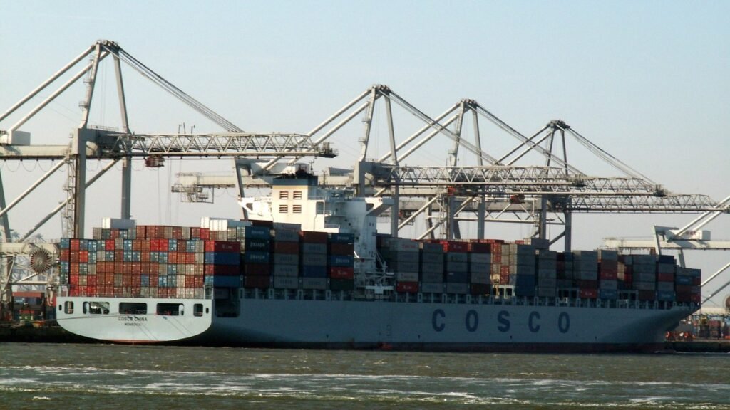 बंदरगाह पर क्रेनों के बीच कंटेनरों से लदा चीनी शिपिंग कंपनी COSCO का मालवाहक जहाज
