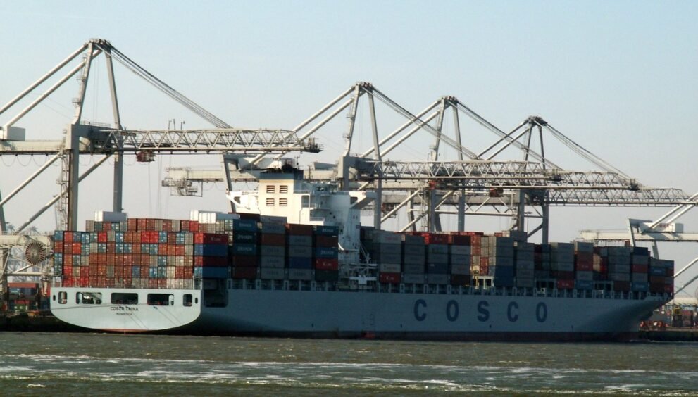 बंदरगाह पर क्रेनों के बीच कंटेनरों से लदा चीनी शिपिंग कंपनी COSCO का मालवाहक जहाज