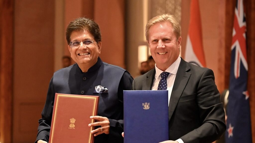 भारत और न्यूजीलैंड के झंडों के सामने मुक्त व्यापार समझौते के दस्तावेज थामे पीयूष गोयल और टॉड मैक्ले