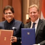भारत और न्यूजीलैंड के झंडों के सामने मुक्त व्यापार समझौते के दस्तावेज थामे पीयूष गोयल और टॉड मैक्ले