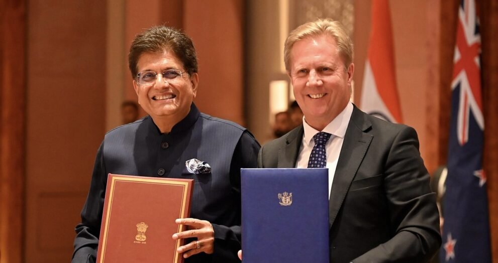 भारत और न्यूजीलैंड के झंडों के सामने मुक्त व्यापार समझौते के दस्तावेज थामे पीयूष गोयल और टॉड मैक्ले