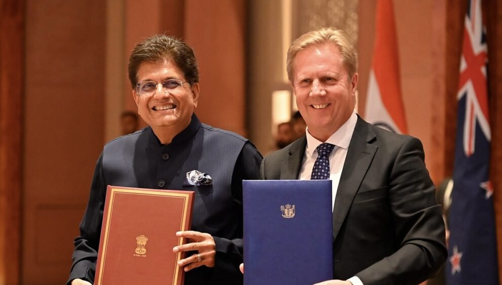 भारत और न्यूजीलैंड के झंडों के सामने मुक्त व्यापार समझौते के दस्तावेज थामे पीयूष गोयल और टॉड मैक्ले