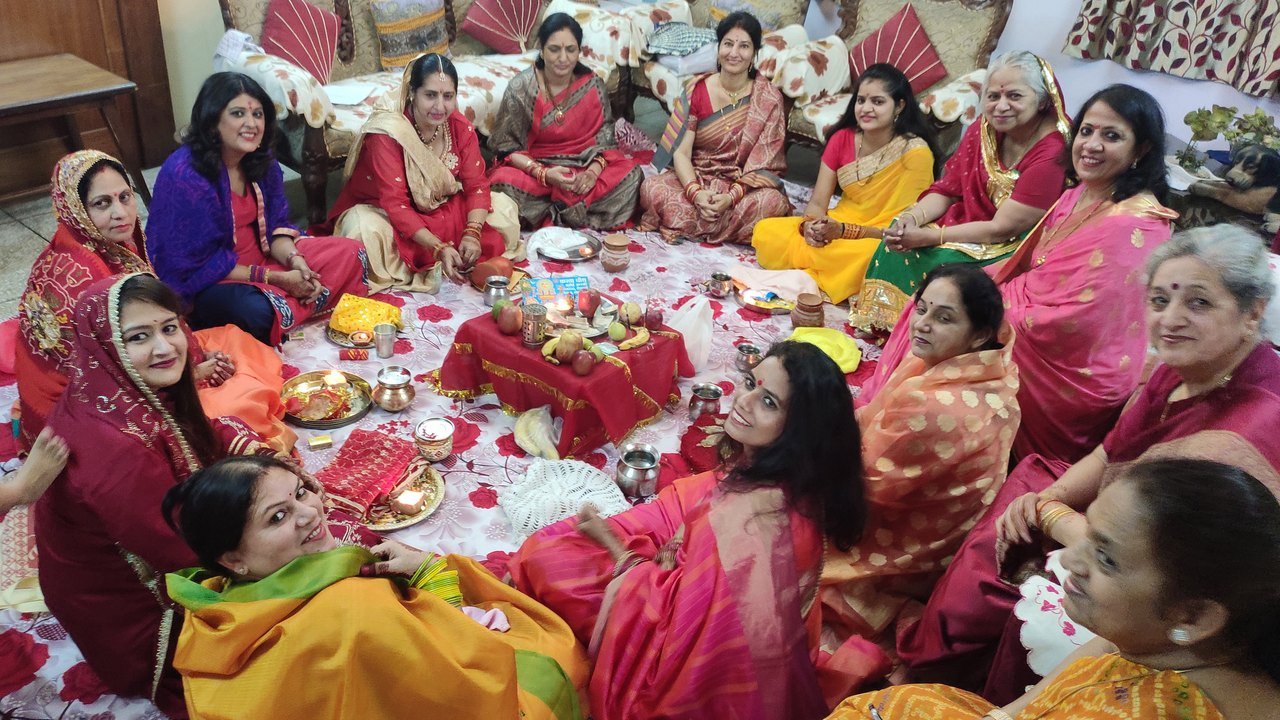 करवा चौथ की पूजा के लिए घेरा बनाकर बैठी महिलाएं, बीच में पूजा की थालियां और करवे